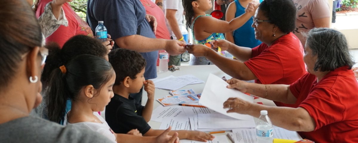 families gather around a table with two women wearing red shirts providing information about hurricane response