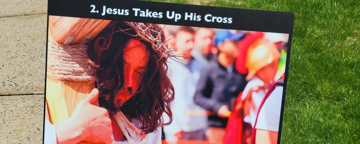 A lawn sign depicting a man dressed as Jesus (long brown hair and a crown of thorns) carrying a large wooden cross on his shoulder. A crowd, blurry in the background, is watching.