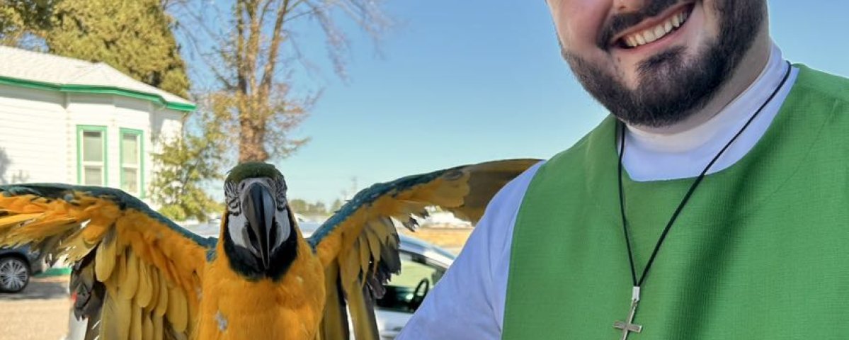 A man in church vestments smiles as he holds a parrot with outstretched wings on his arm.
