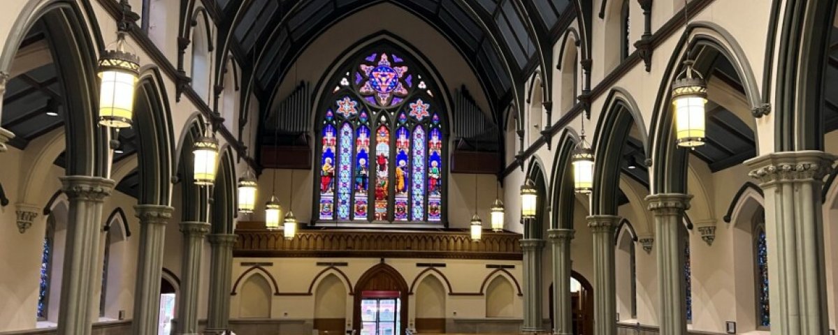 Interior view of Trinity Episcopal Cathedral in Pittsburgh, with rows of wooden pews, tall Gothic arches and columns, hanging lantern lights, and a large stained glass window above the main entrance.