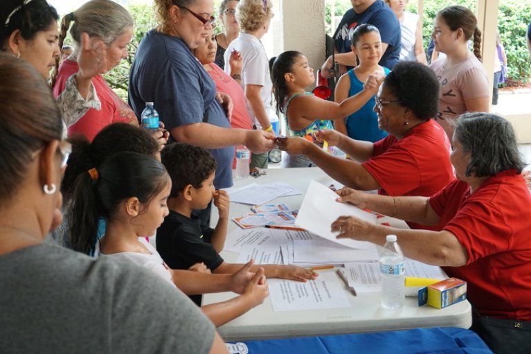 families gather around a table with two women wearing red shirts providing information about hurricane response