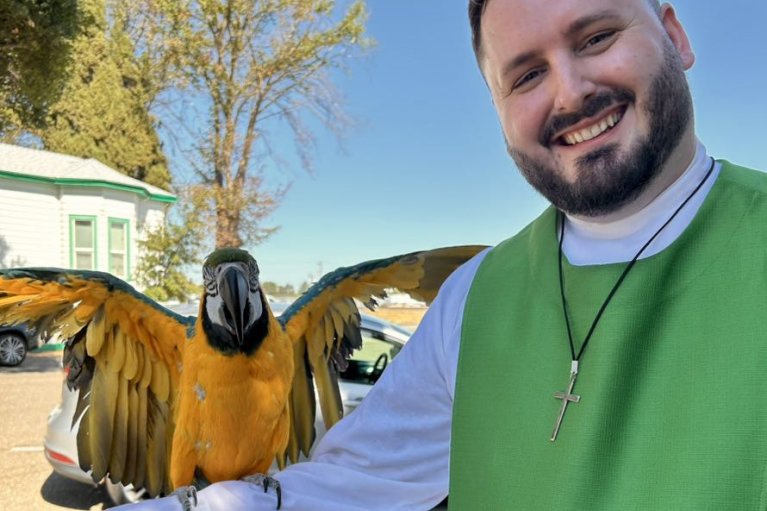 A man in church vestments smiles as he holds a parrot with outstretched wings on his arm.