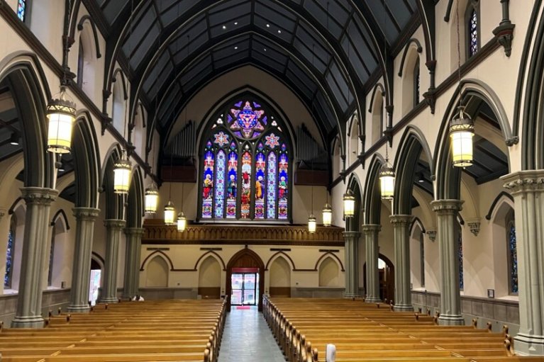 Interior view of Trinity Episcopal Cathedral in Pittsburgh, with rows of wooden pews, tall Gothic arches and columns, hanging lantern lights, and a large stained glass window above the main entrance.