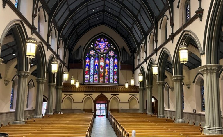 Interior view of Trinity Episcopal Cathedral in Pittsburgh, with rows of wooden pews, tall Gothic arches and columns, hanging lantern lights, and a large stained glass window above the main entrance.